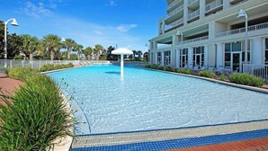 Una piscina amb un parasol blanc, envoltada d'un edifici amb balcons i un cel blau clar.