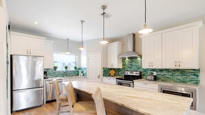 Una cocina moderna con armarios blancos, un refrigerador de acero inoxidable, una isla grande con taburetes y luminarias colgantes.