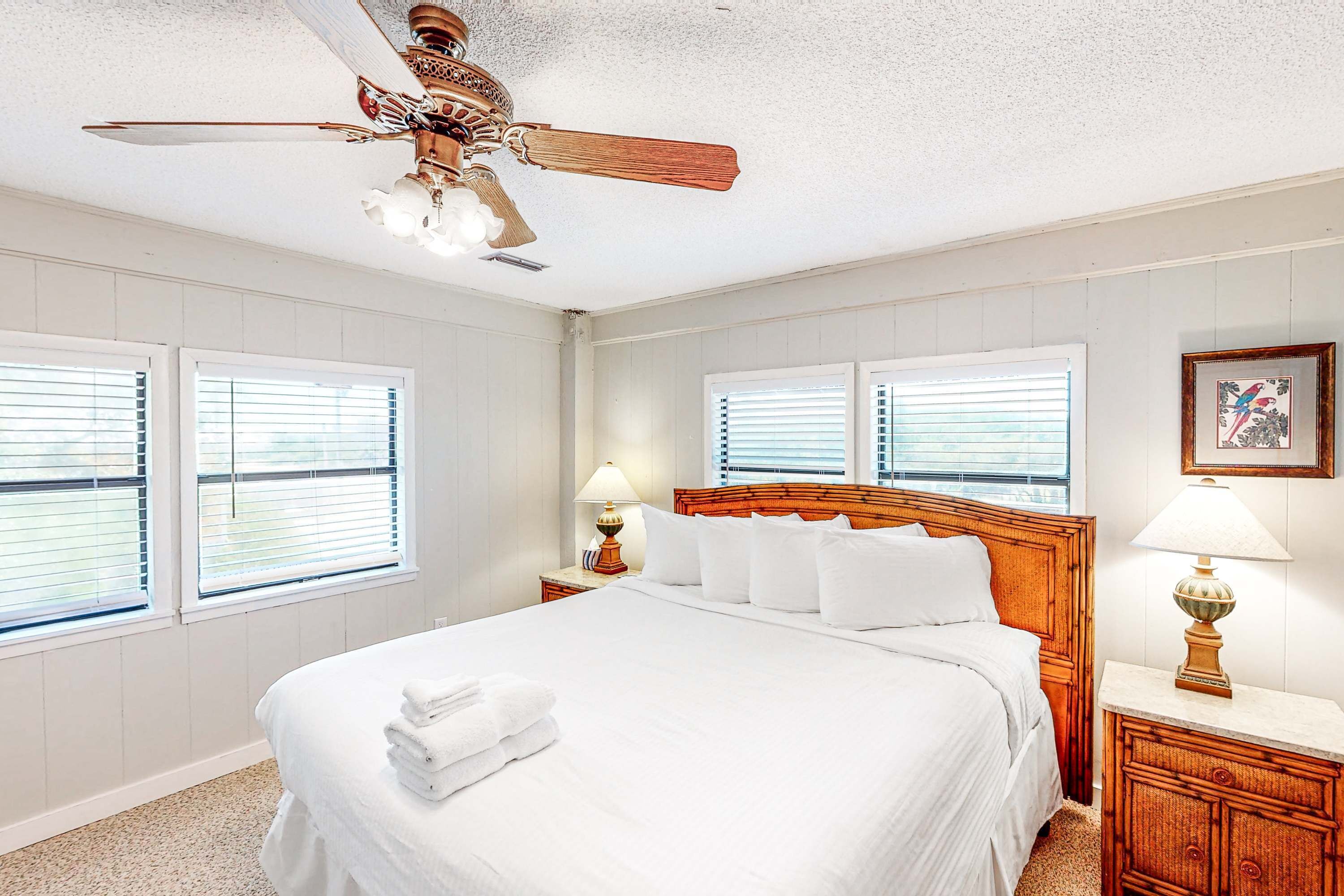 A neatly arranged bedroom with a large bed, two bedside tables with lamps, a ceiling fan, and three windows with blinds.