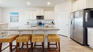A kitchen with white cabinets, a stainless steel refrigerator, a granite countertop, and three woven stools. - Spacious home steps from the beach with fast WiFi & washer/dryer (Port St. Joe)