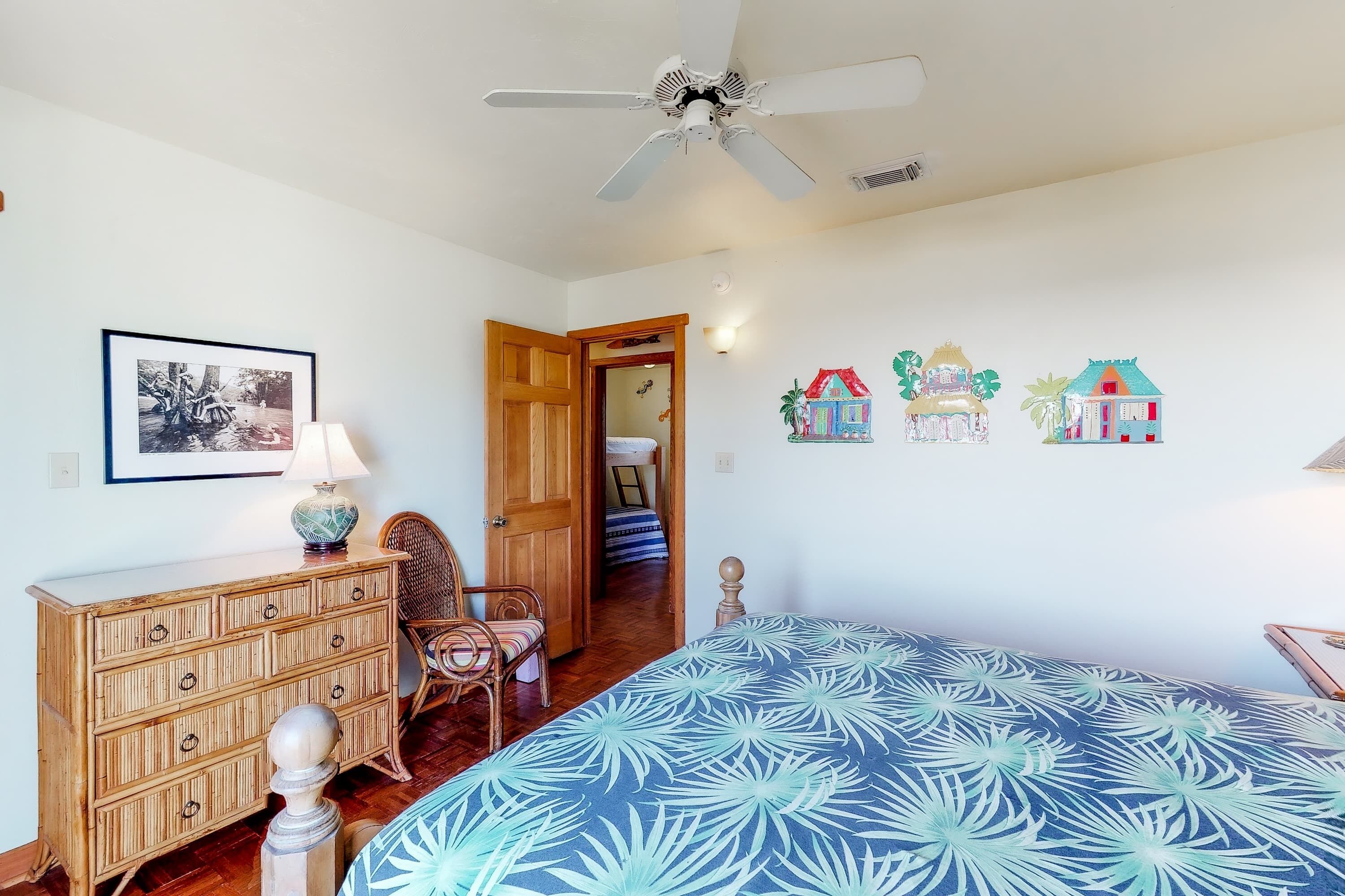 Une chambre à coucher comprenant un lit, une commode en bois, une chaise, un ventilateur de plafond et une porte donnant sur une autre pièce.