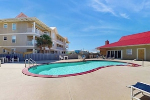 A swimming pool with lounge chairs and a fence, surrounded by buildings with balconies. - Cozy beach condo with amazing views, beach access & on-site pool (Navarre Beach)