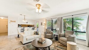 Amplia sala de estar con un sofá blanco, una mesita redonda y un sillón gris. Se ve un área de cocina al fondo, y la habitación tiene ventanas grandes con persianas.