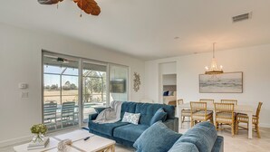 A spacious living room with a blue sofa, a dining area with wicker chairs, and a large window offering a view of the outdoors. - Golfers Dream Ibis Landing Golf (Lehigh Acres)