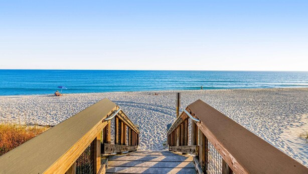 Une promenade en bois mène à une plage de sable fin, sous un ciel bleu et dégagé.