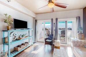 Un salón espacioso con un televisor en un estante azul, ventilador de techo, balcón con vistas y un área de estar confortable.