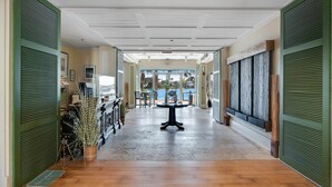 Un couloir d’hôtel avec une table centrale, des volets verts et une vue sur l’extérieur.