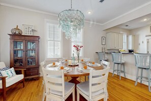 A dining area with a round table set for four, surrounded by white chairs. A chandelier hangs from the ceiling, and a cabinet with glass doors is visible to the left. - Coastal home with 2 community pools, bikes, front porch & balcony - near beach (Santa Rosa Beach)