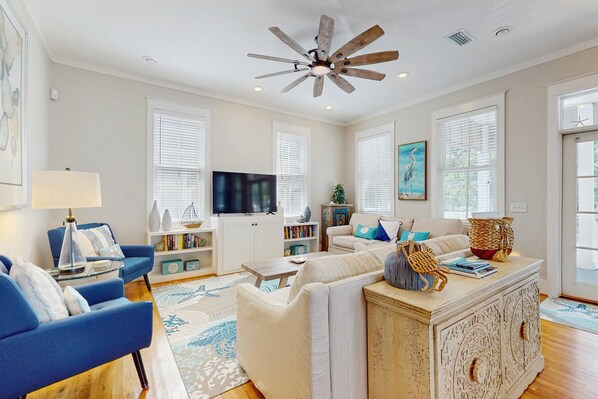 A spacious living room with a ceiling fan, a television, and a large window. - Coastal home with 2 community pools, bikes, front porch & balcony - near beach (Santa Rosa Beach)