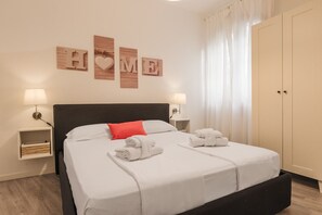 A neatly made bed with white linens and two rolled towels, a red pillow, and a black headboard. There are two bedside tables with lamps, a wall-mounted sign spelling "HOME," and a large wardrobe.