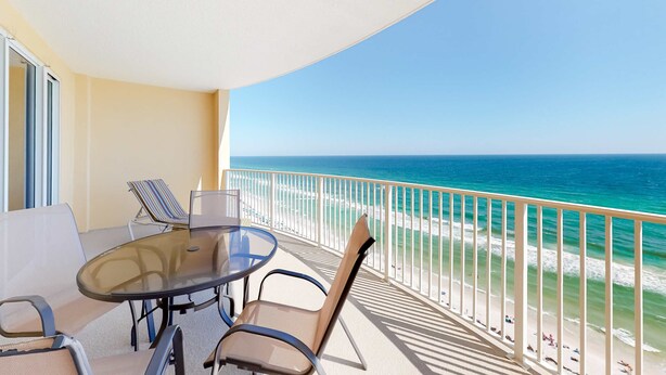 Een balkon met een glazen tafel en stoelen, met uitzicht op een strand met helderblauw water en een zandstrand.