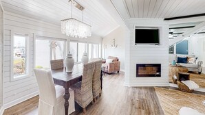Ein geräumiges Wohnzimmer mit Holztisch, Korbstühlen, einer Kristallleuchtre, einem Kamin und einem an der Wand befestigten Fernseher.