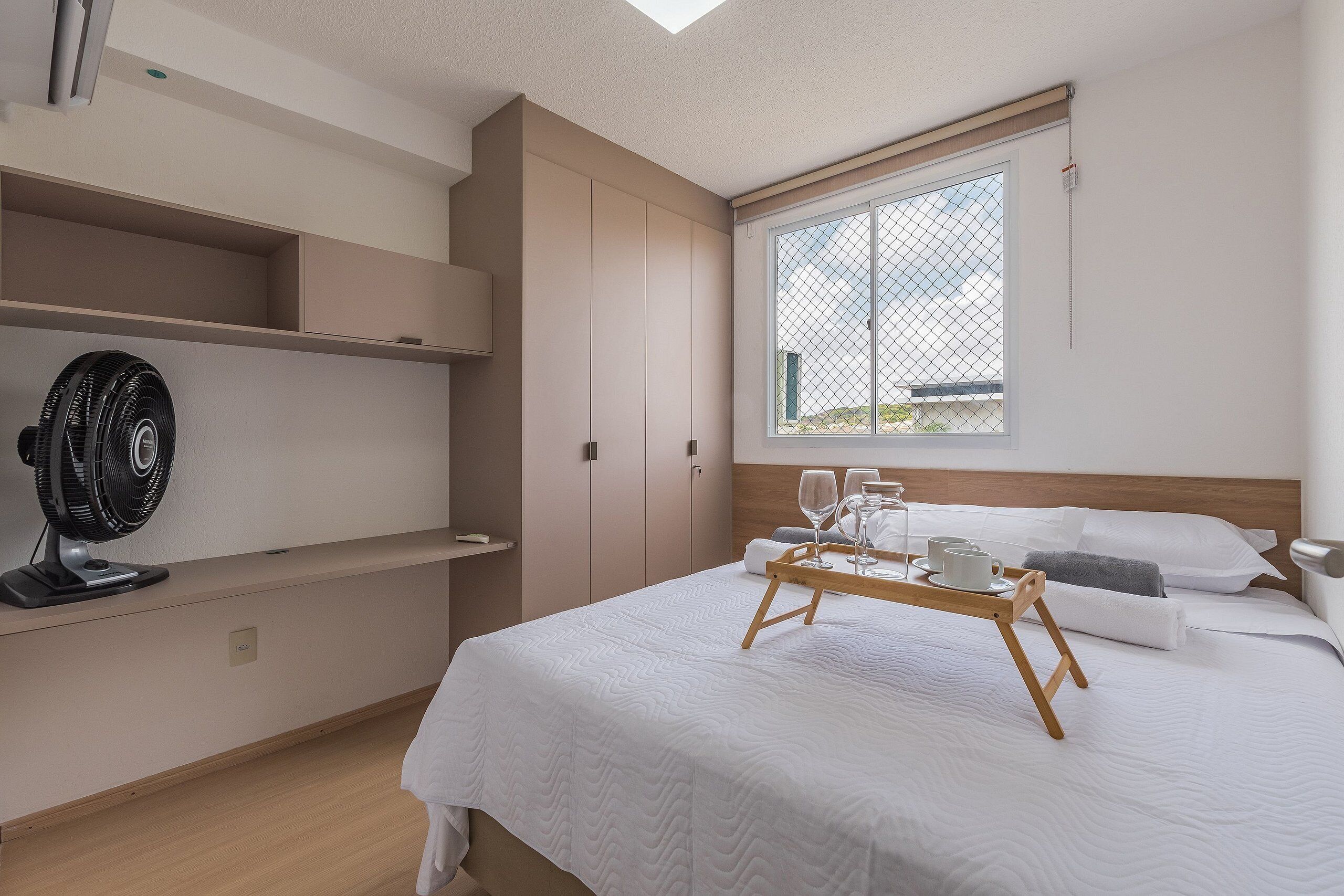 Habitación de hotel con cama, mesita de noche con vasos y bandeja, un ventilador y ventana con vistas.