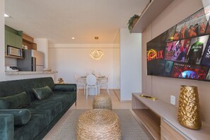 Ein modernes Wohnzimmer mit einem grünen Sofa, einem Esstisch mit Stühlen, einem an der Wand montierten Fernseher und einer Küchenzeile mit Mikrowelle und Kühlschrank.