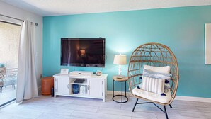 A room with a teal wall, a flat-screen TV, a white console table, a wicker chair, and a small table with a lamp. - Relaxing townhome with pool, tennis court, beach access, balcony, patio, & TV (Panama City Beach)