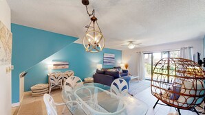 A modern living room with a glass dining table, white chairs, a blue sofa, and a hanging spherical light fixture. - Relaxing townhome with pool, tennis court, beach access, balcony, patio, & TV (Panama City Beach)