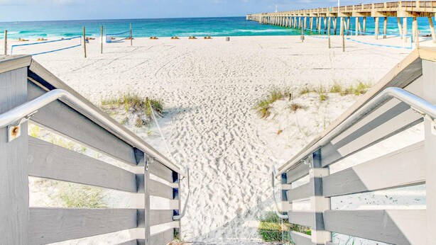 A beach with a wooden staircase leading down to the sand, a pier extending into the ocean, and a clear sky. - Relaxing townhome with pool, tennis court, beach access, balcony, patio, & TV (Panama City Beach)