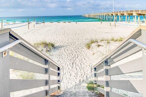 Een strand met een houten trap die naar het zand leidt, een steiger die zich uitstrekt over de oceaan en een heldere lucht.