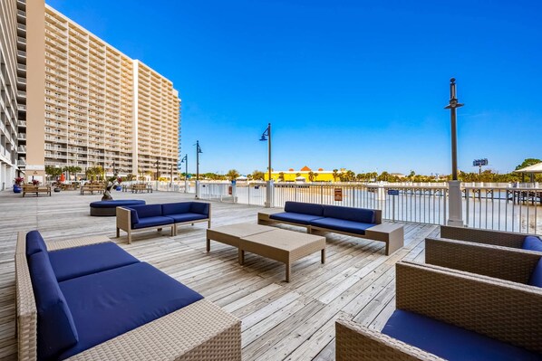 A wooden deck with blue cushioned seating, a railing, and a large building in the background. - Spacious condo with bunk beds, rooftop pool, hot tub, washer/dryer & central AC (Panama City Beach)