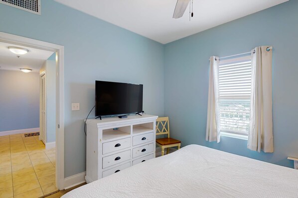 A bedroom with a bed, a TV on a dresser, a chair, and a window with curtains. - Spacious condo with bunk beds, rooftop pool, hot tub, washer/dryer & central AC (Panama City Beach)