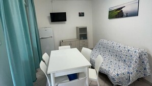 Ett kompakt vardagsrum med ett matbord, vita stolar, en soffa med ett blommigt överdrag, en platt-TV och ett litet kylskåp.