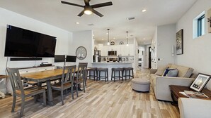 A modern kitchen with white cabinets, a central island with stools, and a dining area with a wooden table and chairs. - Gorgeous coastal home, dog friendly, private hot tub & close to beach (Panama City Beach)