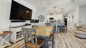 A modern kitchen with white cabinets, a large flat-screen TV, and a wooden dining table with chairs. - Gorgeous coastal home, dog friendly, private hot tub & close to beach (Panama City Beach)