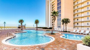 A swimming pool with a tiled deck, surrounded by lounge chairs and palm trees, adjacent to a multi-story building with balconies. - Gulf View & Laketown Wharf Condo with full kitchen, shared pool and beach access (Panama City Beach)