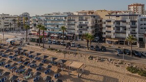 Uma área urbana costeira com edifícios de apartamentos modernos, uma praia de areia com espreguiçadeiras e um céu claro.