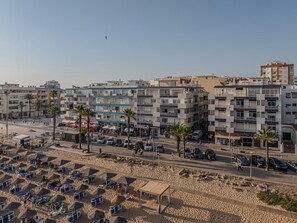 Ein Küstenort mit modernen Wohngebäuden, einem Sandstrand mit Liegestühlen und blauem Himmel.