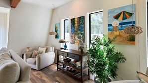 Ein Wohnzimmer mit einer Couch, einem Sessel und einem Holztisch mit einer Lampe. An der Wand hängen Bilder und es steht ein Blumentopf dort.