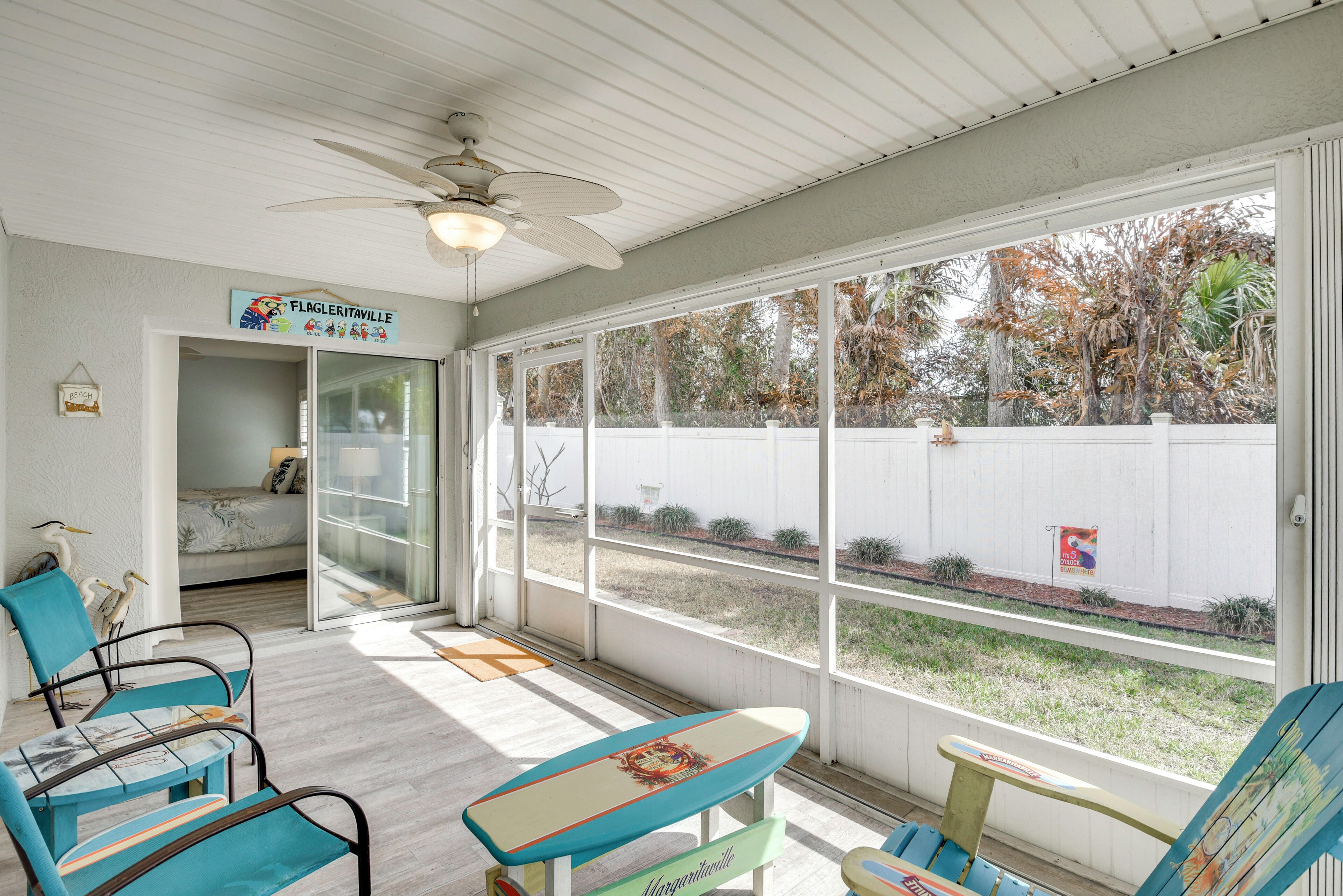 A sunlit room with a ceiling fan, a bed, and a view of a white fence and trees.