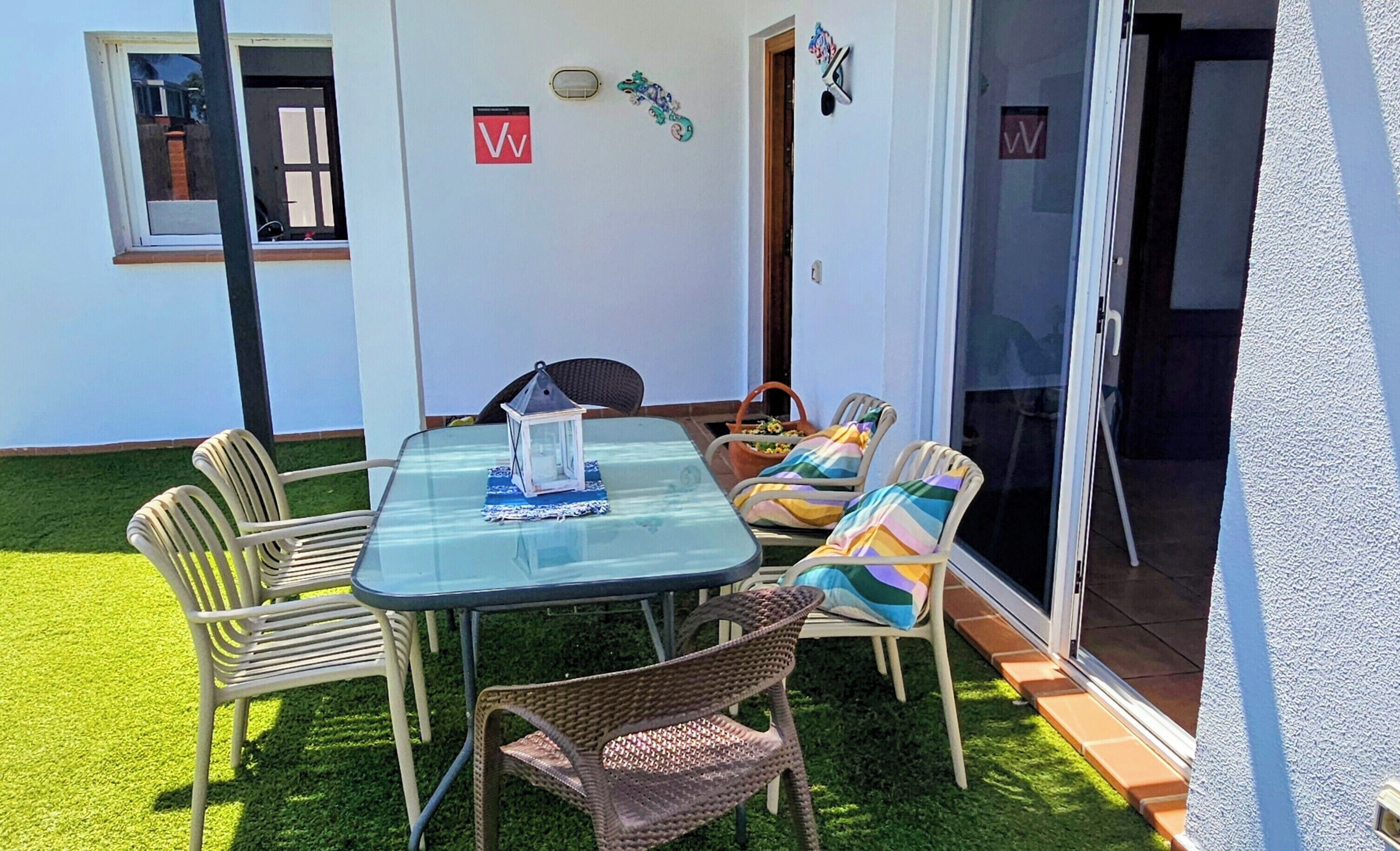 Eine Terrasse im Freien mit einem Glastisch, Korbstühlen, einer Tür mit einem 'W'-Schild und einem kleinen Garten.