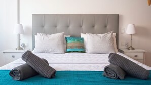 Un lit bien fait, avec une tête de lit grise, des oreillers blancs et turquoise, et des serviettes grises.