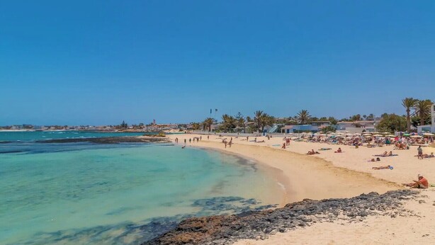 Une plage aux eaux turquoise et limpides, avec un rivage sablonneux et quelques bâtiments visibles au loin.