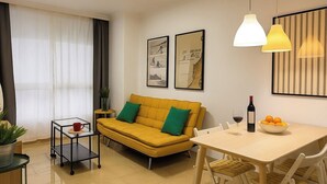 A modern living room with a yellow sofa, a dining table with chairs, a small side table, and a potted plant. - Miguel Rosas (Las Palmas de Gran Canaria)
