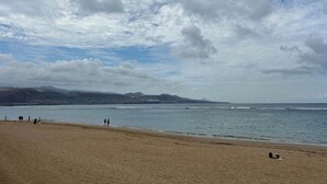 A beach with people walking along the shore, a clear sky, and distant mountains. - Miguel Rosas (Las Palmas de Gran Canaria)