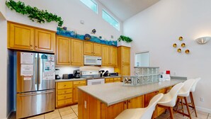 A modern kitchen with wooden cabinets, a stainless steel refrigerator, a microwave, and a large island with bar stools. - Lagoon-front home with dock & soaking bathtub (Panama City Beach)