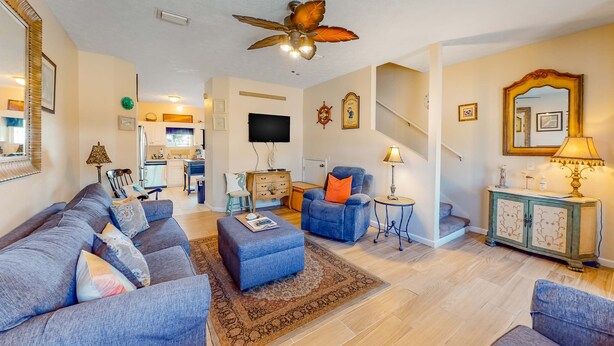 Een ruime woonkamer met een grote bank, een blauwe fauteuil, een klein voetenbankje, een plafondventilator, een trap en een keuken op de achtergrond.