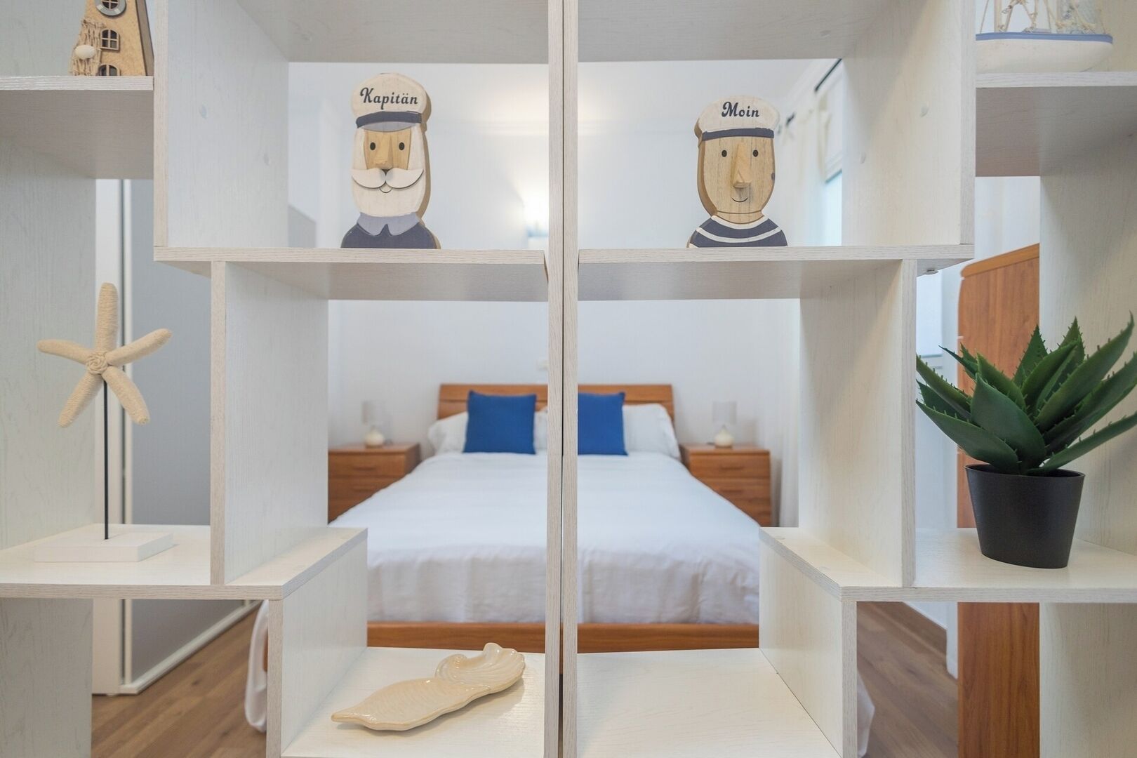 Habitación de hotel con cama, dos mesitas de noche, una decoración con forma de estrella de mar, una planta en maceta y dos figuras de madera etiquetadas como "Kapitan" y "Moin".