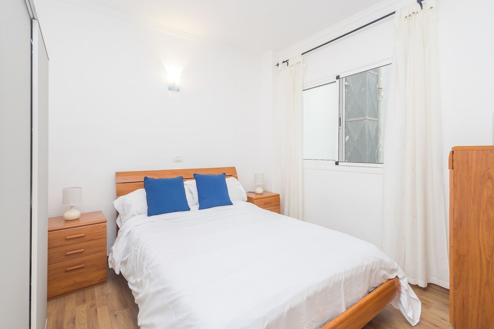 Une chambre à coucher avec un lit en bois, une literie blanche, deux tables de chevet avec des lampes, une fenêtre avec des rideaux et une armoire en bois.