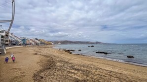A beach with a curved walkway, buildings along the shore, and rocky outcrops in the water. - Estudio en primera línea de playa (Las Palmas de Gran Canaria)