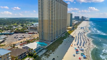 A high-rise hotel with a beach view, a parking lot, and a long stretch of beach with umbrellas and people.