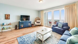 A living room with a sofa, a coffee table, a TV, and a balcony with curtains. - 14th-Floor Condo w/Gulf Views, W/D, Shared Pools, Hot Tubs, WiFi, & Central A/C (Panama City Beach)