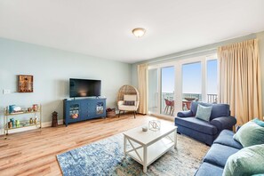 A living room with a sofa, a coffee table, a TV, and a balcony with curtains. - 14th-Floor Condo w/Gulf Views, W/D, Shared Pools, Hot Tubs, WiFi, & Central A/C (Panama City Beach)