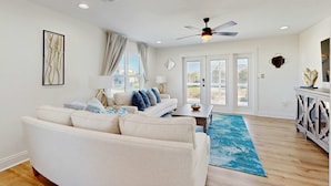A spacious living room with a beige sofa, a coffee table, and a blue rug. There is a ceiling fan, a window with curtains, and a door leading outside. - Nautical home in beach community with Water views close to beach access (Navarre)