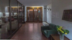 Um lobby de hotel com uma escadaria que leva ao andar superior, um sofá verde, um vaso com flores amarelas e um painel de vidro.