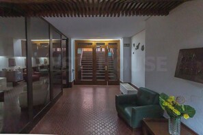 Un vestíbulo de hotel con una escalera que lleva al piso superior, un sofá verde, un jarrón con flores amarillas y una mampara de vidrio.