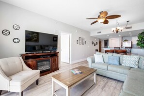 A living room with a sofa, armchair, coffee table, TV, and ceiling fan. - Beachfront condo on 10th floor w/shared pools (4), hot tub, tennis - great views (Panama City Beach)