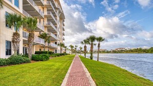 A waterfront hotel with a brick pathway leading to the water, surrounded by palm trees and a well-maintained lawn. - Sunset-view dog-friendly condo w balcony overlooking water, beach access, pool & hot tub (Panama City Beach)